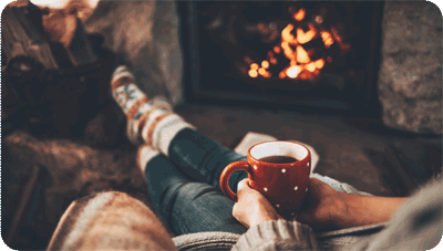 A person sitting on a couch, legs crossed, holding a warm cup in front of a cozy fireplace